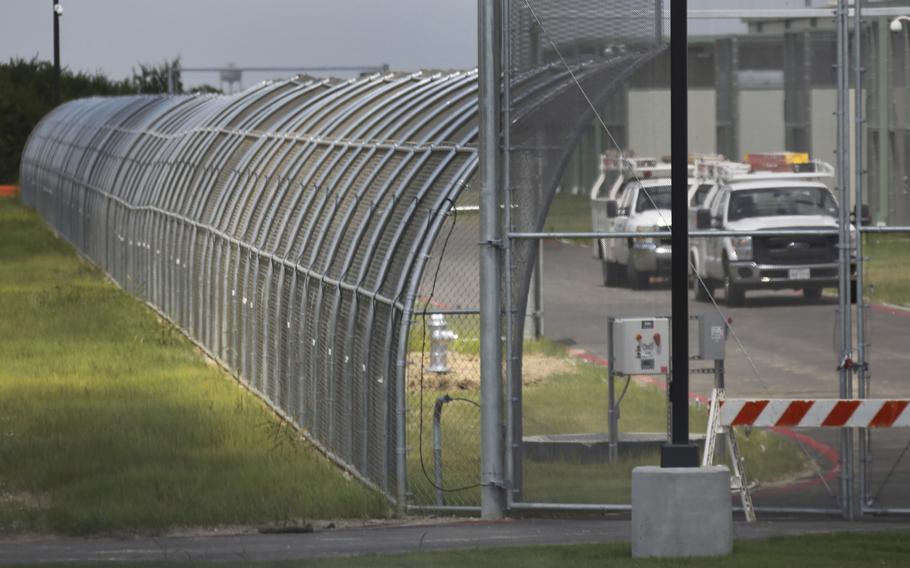 Two white trucks drive on a road behind the chain link fence of a government detention facility.