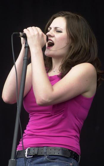 A singer in a purple shirt holds her microphone to her mouth with both hands.