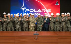 A group of South Korean and U.S. military personnel, all wearing camouflage uniforms and white caps, pose and applaud. Two senior officers, one from each country, are shaking hands at the center in front of a large screen displaying the “POLARIS LYNX” logo, which features a stylized star and a planet-like background. 