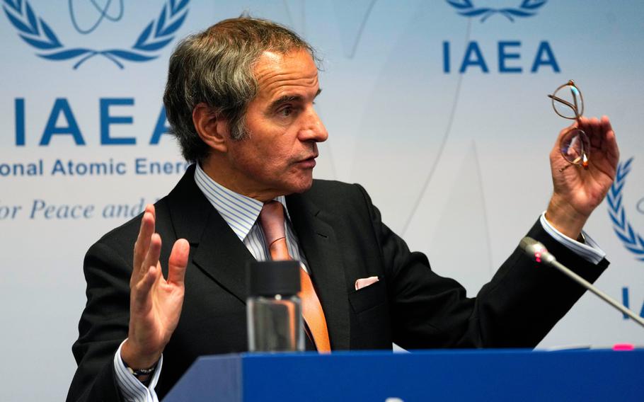 A man in a suit gestures with his arms spread while standing at a podium and speaking into a microphone in front of a white wall reading “IAEA.”