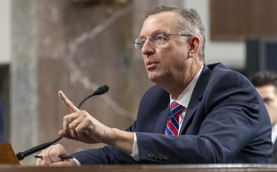 Collins seated at a table, speaking into a microphone, pointing the index finger on his left hand and holding a pen in his right hand.