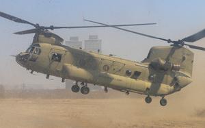 A U.S. Army CH-47 Chinook helicopter in tan desert camouflage hovers low over sandy ground, kicking up a large dust cloud. T