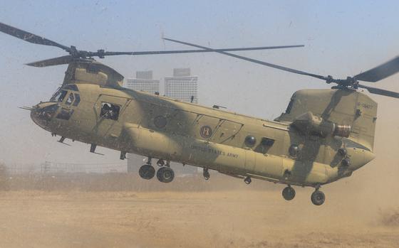 A U.S. Army CH-47 Chinook helicopter in tan desert camouflage hovers low over sandy ground, kicking up a large dust cloud. T