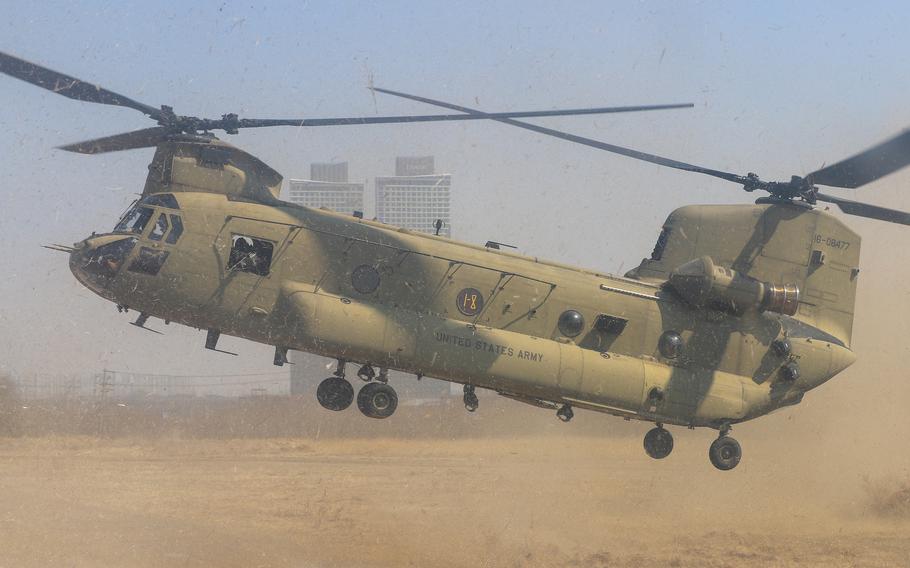 A U.S. Army CH-47 Chinook helicopter in tan desert camouflage hovers low over sandy ground, kicking up a large dust cloud. 