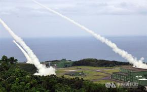 Multiple missiles launch from a coastal military base, leaving white smoke trails as they ascend over the ocean. The installation features green buildings surrounded by hills and vegetation. A CNA watermark visible in corner.
