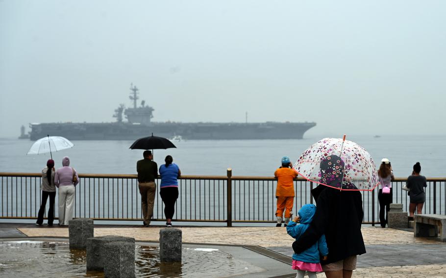 People holding umbrellas watch from the shore as an aircraft carrier sails away.