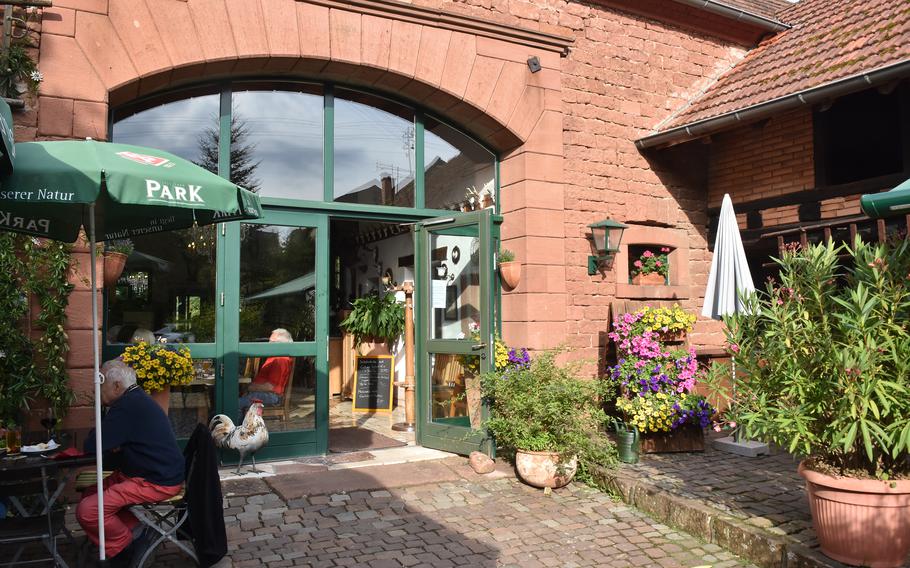 The exterior of a restaurant housed in a charming red sandstone building with green-framed windows and doors. The entrance features an arched doorway with green awnings, and the courtyard is decorated with colorful flowering plants in terracotta pots.