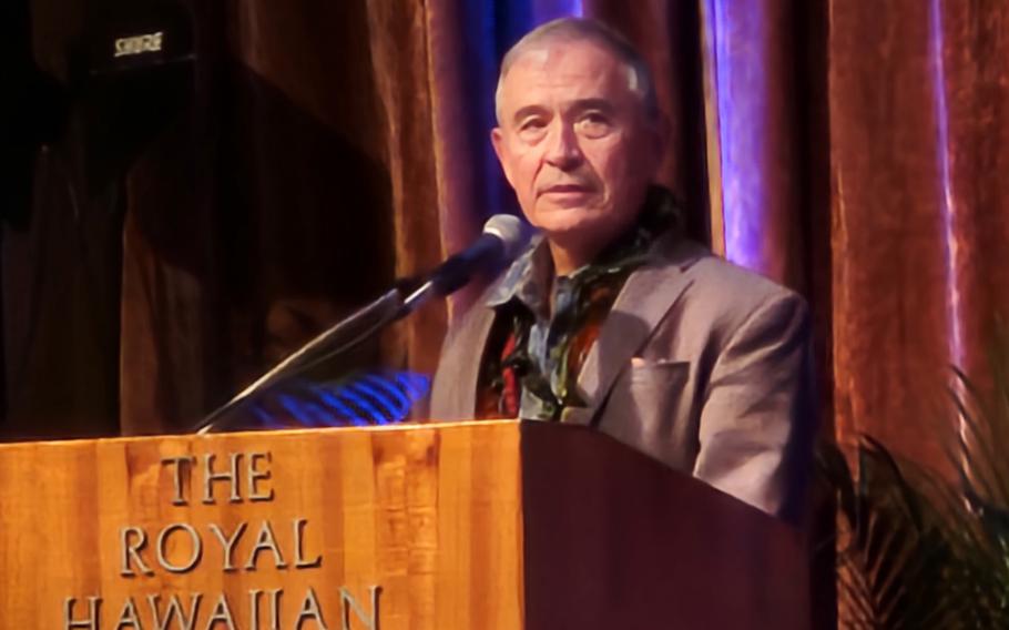 A man in a brown blazer stands at a podium and speaks into a microphone.