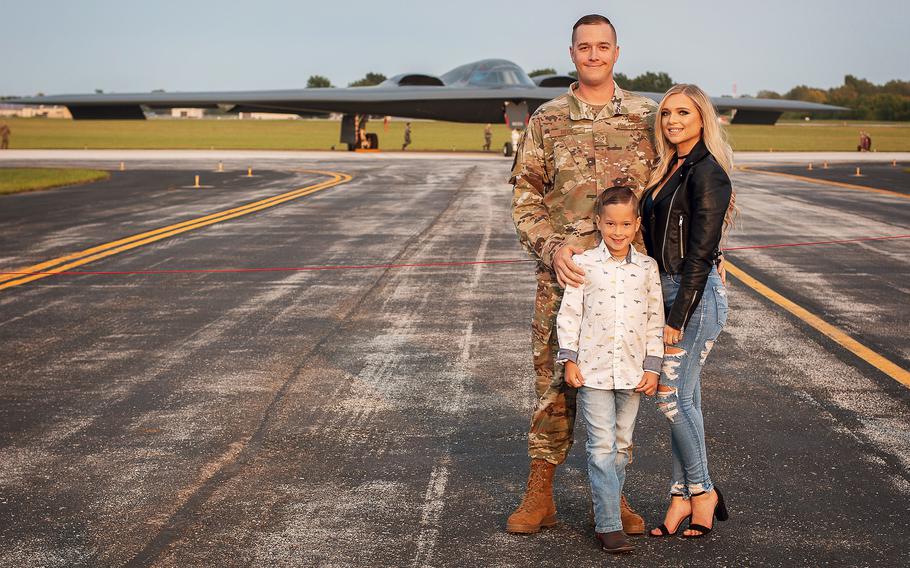 A man in a camouflage uniform poses with a woman and a boy on a military runway with a fighter jet in the background.