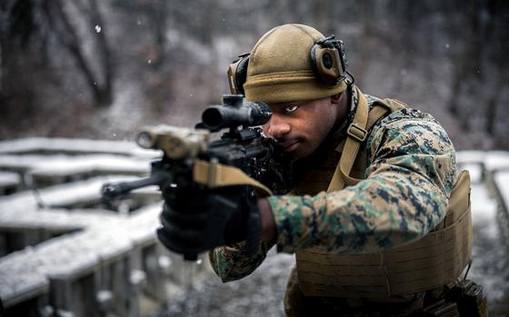 A marine in camouflage uniform and tactical gear aims a rifle with an optical scope in a snowy outdoor environment. The person is wearing a tan helmet with mounted equipment, protective eyewear and a tactical vest. .