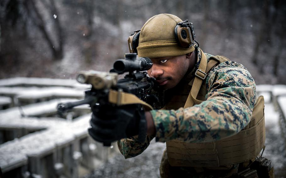 A marine in camouflage uniform and tactical gear aims a rifle with an optical scope in a snowy outdoor environment. The person is wearing a tan helmet with mounted equipment, protective eyewear and a tactical vest. .
