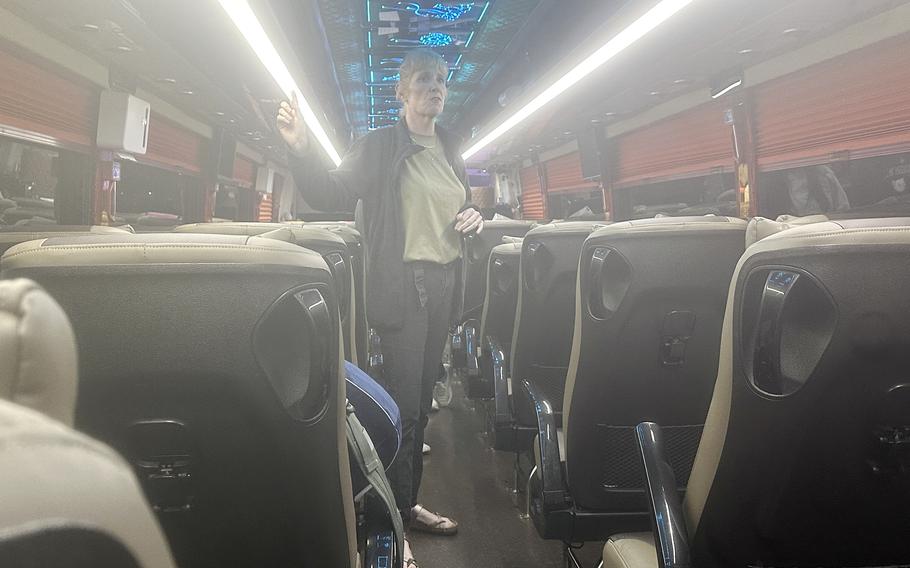 A woman wearing blue jeans and gray shirt stands in the center aisle of a bus and speaks to passengers.
