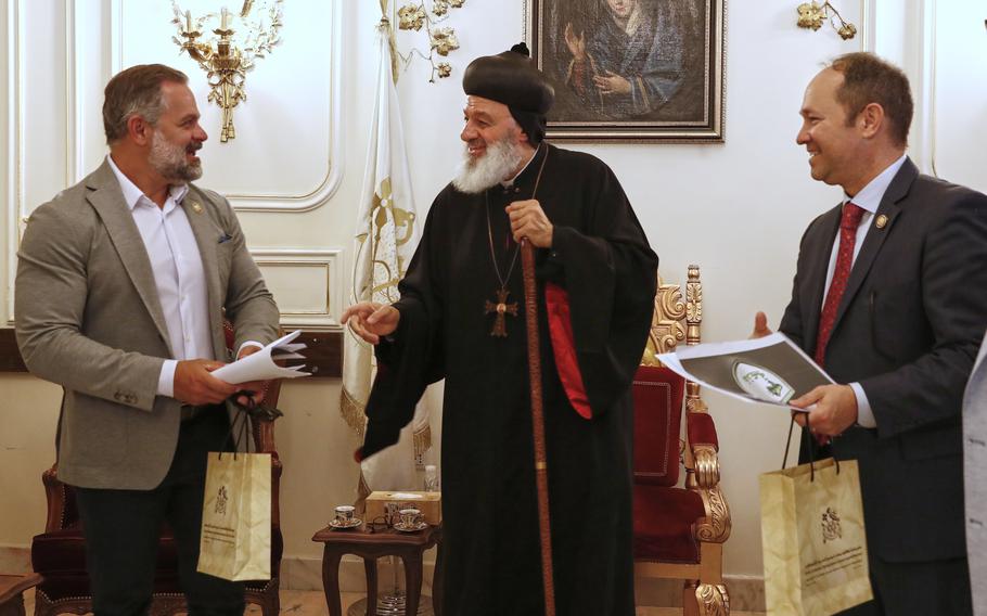 Two congressman speak with a cleric in black robes standing between them in front of a white wall with ornate gold flower decorations.