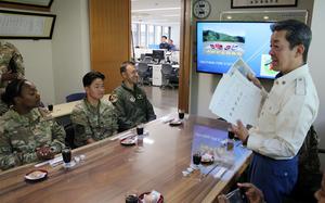 A Japanese fire department official in a white uniform jacket stands addressing seated U.S. Air Force personnel in a conference room. The service members are seated around a wooden table with drinks and small plates. A wall-mounted TV displays “OKUTAMA FIRE STATION” with mountain scenery. 