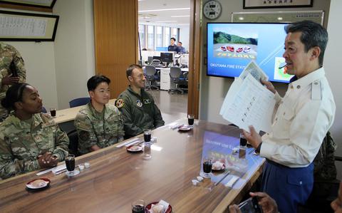 A Japanese fire department official in a white uniform jacket stands addressing seated U.S. Air Force personnel in a conference room. The service members are seated around a wooden table with drinks and small plates. A wall-mounted TV displays “OKUTAMA FIRE STATION” with mountain scenery. 