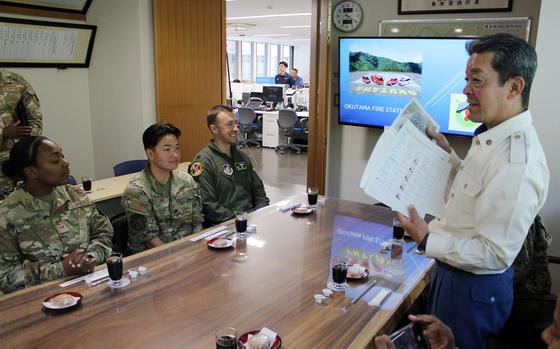 A Japanese fire department official in a white uniform jacket stands addressing seated U.S. Air Force personnel in a conference room. The service members are seated around a wooden table with drinks and small plates. A wall-mounted TV displays “OKUTAMA FIRE STATION” with mountain scenery. 