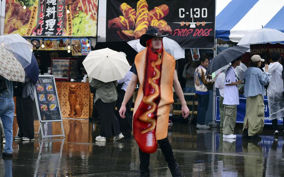 An airman wears a hot dog costume and a cowboy hat in front of festival food booths, with a sign reading “C-130 Prop Dogs” in the background.