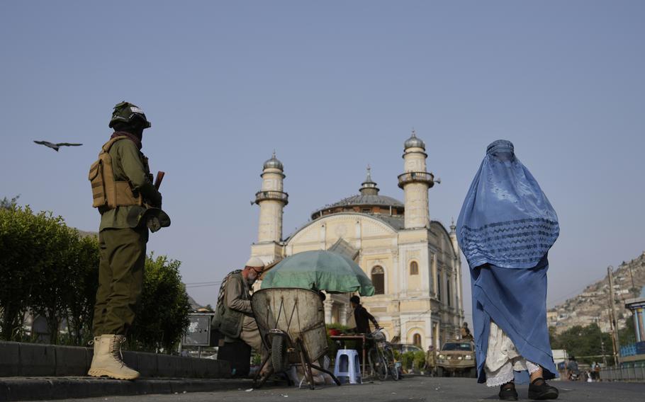 A woman in a blue burqa walking past an armed soldier in military fatigues on a street. In the background is a prominent mosque with white stone architecture; there are also local vendors with carts and umbrellas conducting business on the street. 