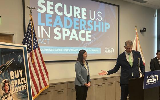 Two people stand before a screen that reads “Secure U.S. leadership in space.”