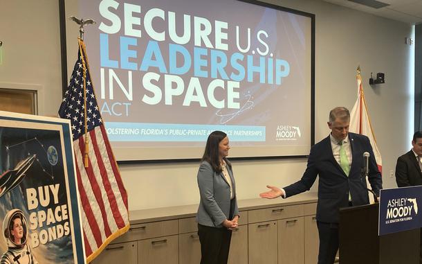 Two people stand before a screen that reads “Secure U.S. leadership in space.”
