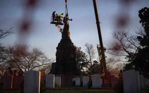 Construction workers on a platform supported by a crane help remove a statue depicting a bronze woman from a large stone pedastal, with rows of white grave markers in the foreground.