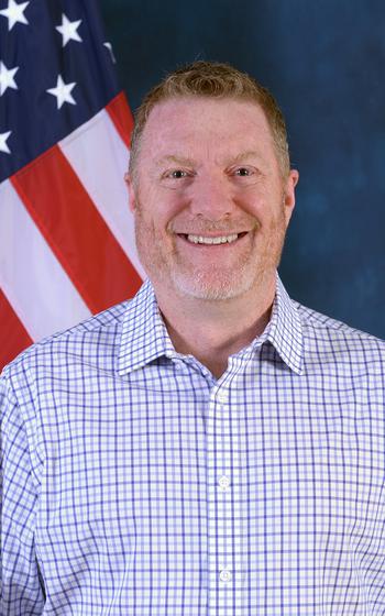 Trubey smiling and wearing a checkered shirt, standing with an American flag behind his right shoulder.