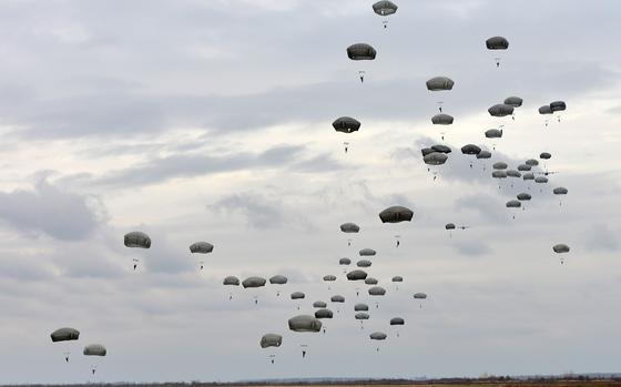 Dozens of military paratroopers descending with deployed parachutes against a cloudy gray sky.