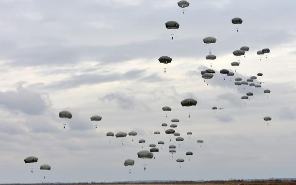 Dozens of military paratroopers descending with deployed parachutes against a cloudy gray sky.