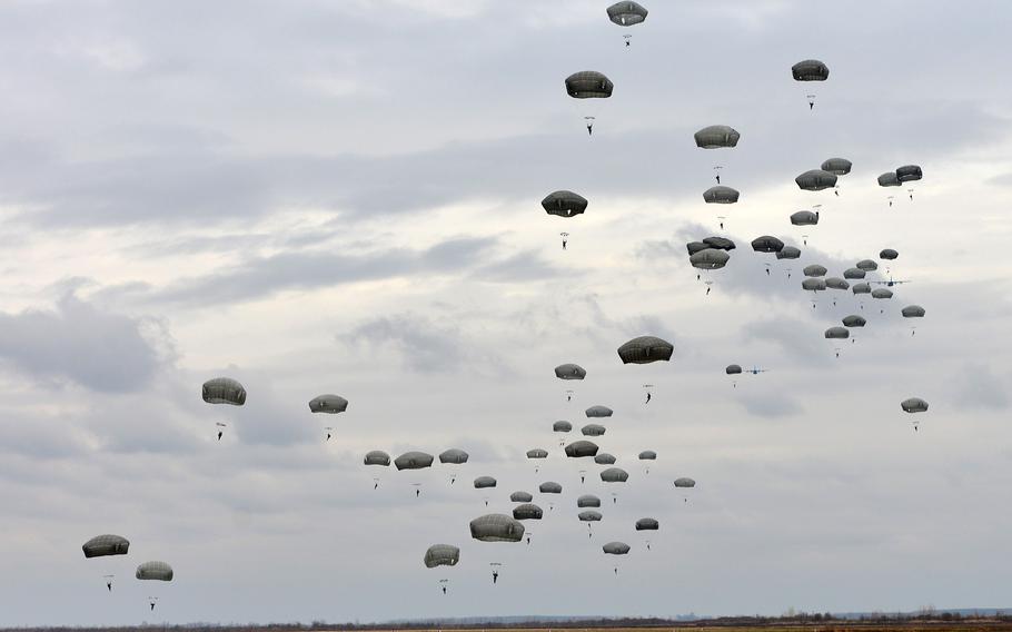 Dozens of military paratroopers descending with deployed parachutes against a cloudy gray sky.