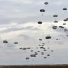 Dozens of military paratroopers descending with deployed parachutes against a cloudy gray sky.