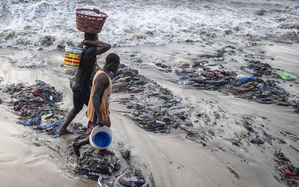 Fishermen walk over piles of trash and discarded textile waste on the shore at Jamestown Harbor in Accra, Ghana. Old clothing collected for transport to developing nations for repurposing, resale or donation often overwhelms the recipients. 