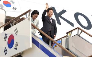 The South Korean president stands and waves at the top of a staircase outside the door to an airplane, while his wife stands behind him.