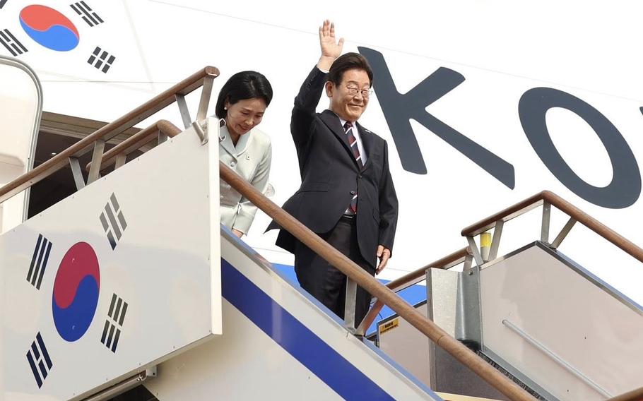 The South Korean president stands and waves at the top of a staircase outside the door to an airplane, while his wife stands behind him.
