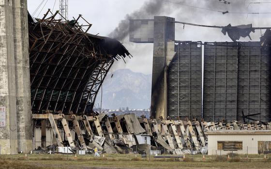 Tustin, CA - November 13: A disaster cleanup crew picks up potentially toxic debris around the still-burning WWII-era blimp hangar at the former air base in Tustin Monday, Nov. 13, 2023. Flare-ups and toxic air from last week's destructive hangar fire in Tustin continue to cause trouble for nearby residents. The City of Tustin took to X, formerly Twitter, to confirm that the western wall of the 17-story building reignited Sunday night. Orange County Fire Authority personnel remained on the scene keeping watch of the blaze on Monday morning, with one firefighter telling KTLA 5's Annie Rose Ramos that all they could do was let it burn out. (Allen J. Schaben / Los Angeles Times)