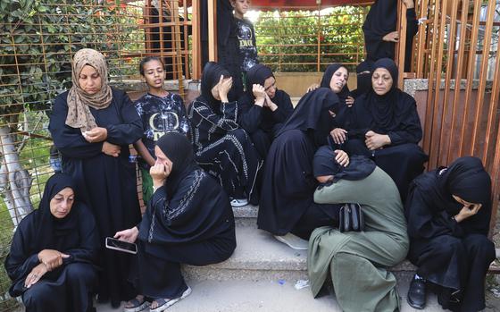 A group of Palestinian women in black robes and head coverings sit on the steps in front of a hospital entrance in various states of mourning.