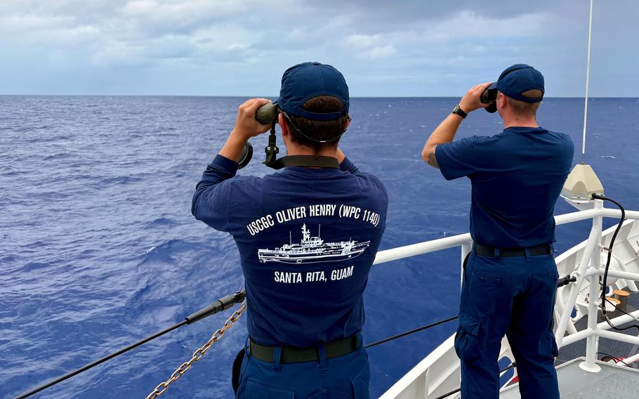 Two Coast Guardsmen, one with a “USCG Oliver Henry” design on the back of his shirt, use binoculars to look toward the water. 