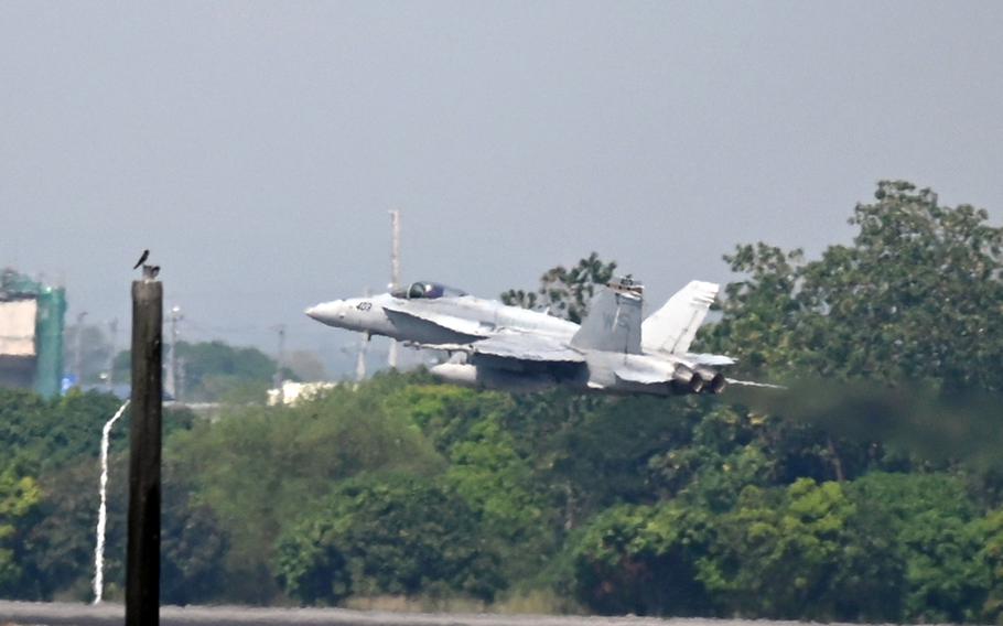 A F/A-18 jet takes off at low altitude over a tree-lined area, with heat distortion visible behind the engines.