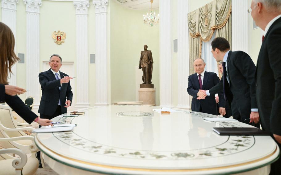 Several people in suits, one of whom is Vladimir Putin, ring around an oval table.