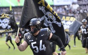 A football player wearing a black uniform with number 54 and “BIVINS” on the nameplate runs onto the field carrying a large black flag featuring a white skull and crossbones design with yellow “ARMY” text. 