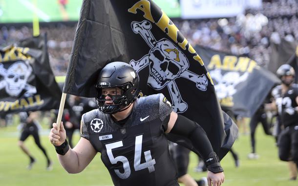 A football player wearing a black uniform with number 54 and “BIVINS” on the nameplate runs onto the field carrying a large black flag featuring a white skull and crossbones design with yellow “ARMY” text. 