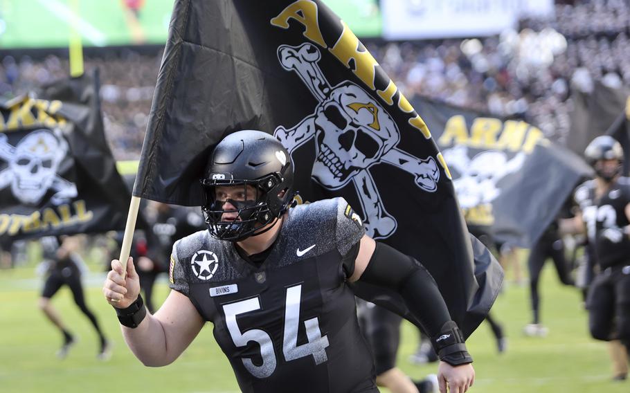 A football player wearing a black uniform with number 54 and “BIVINS” on the nameplate runs onto the field carrying a large black flag featuring a white skull and crossbones design with yellow “ARMY” text. 