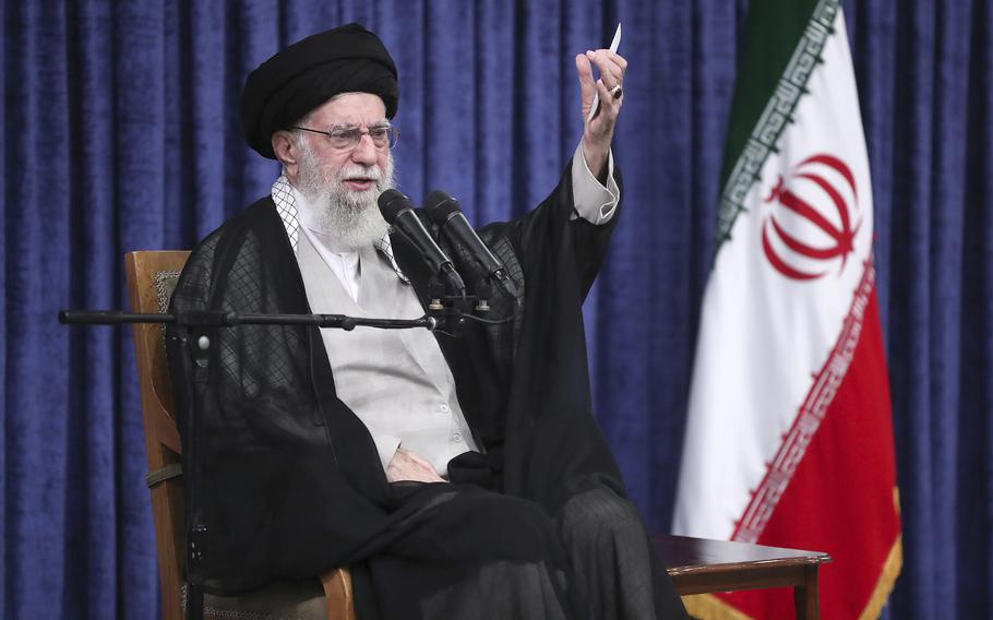 An Iranian man wearing robe, turban and glasses raises his left hand and speaks into two microphones while seated next to an Iranian flag.