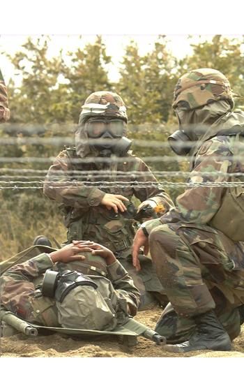 A vertical photo of three soldiers, one lying on the ground and two crouching near the first. All are in gas masks.