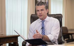 Pete Hegseth speaks while seated at a desk, holding a folder with his right hand and gesturing to it with his left hand.