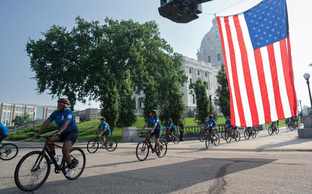 Veterans in blue shirts ride on bikes, with the American flag hanging overhead and a state capitol building in the background.