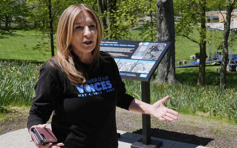 A woman in a black shirt gestures with her arms spread while standing in front of a marker with trees in the background.