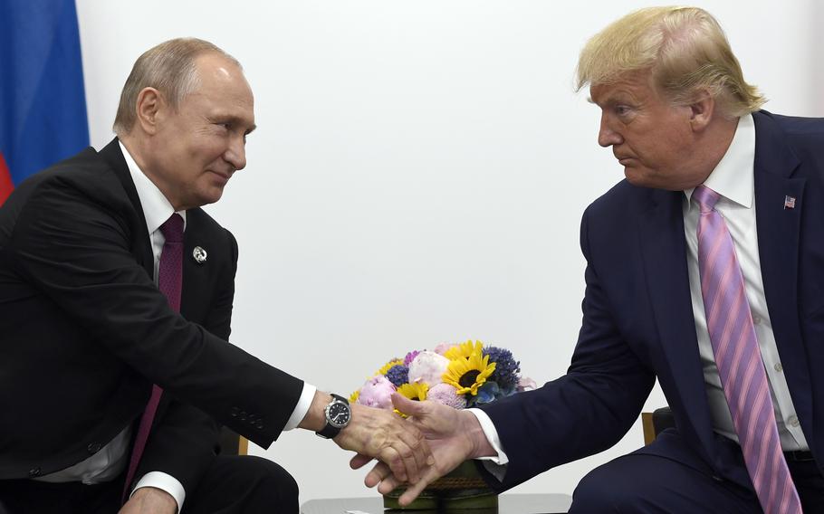 Donald Trump and Vladimir Putin lean towards each other to shake hands in front of a vase of flowers while seated.