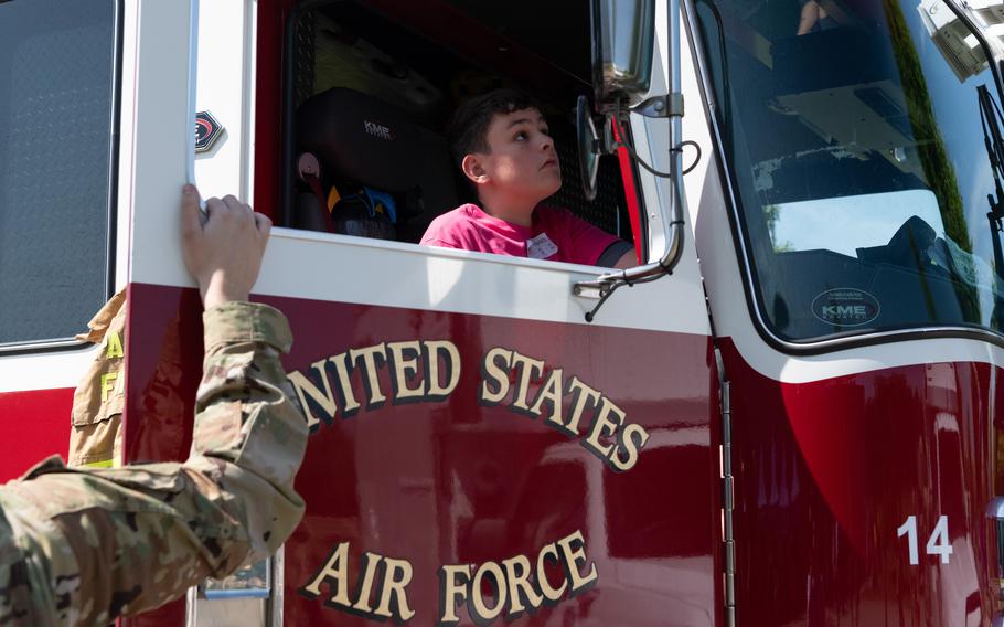 An airman holds the open door of a fire truck as a boy sitting in the passenger seat peers out the front window.