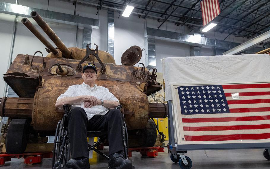 A Marine veteran sits in front of a tank and beside the American flag.