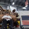 A Marine veteran sits in front of a tank and beside the American flag.